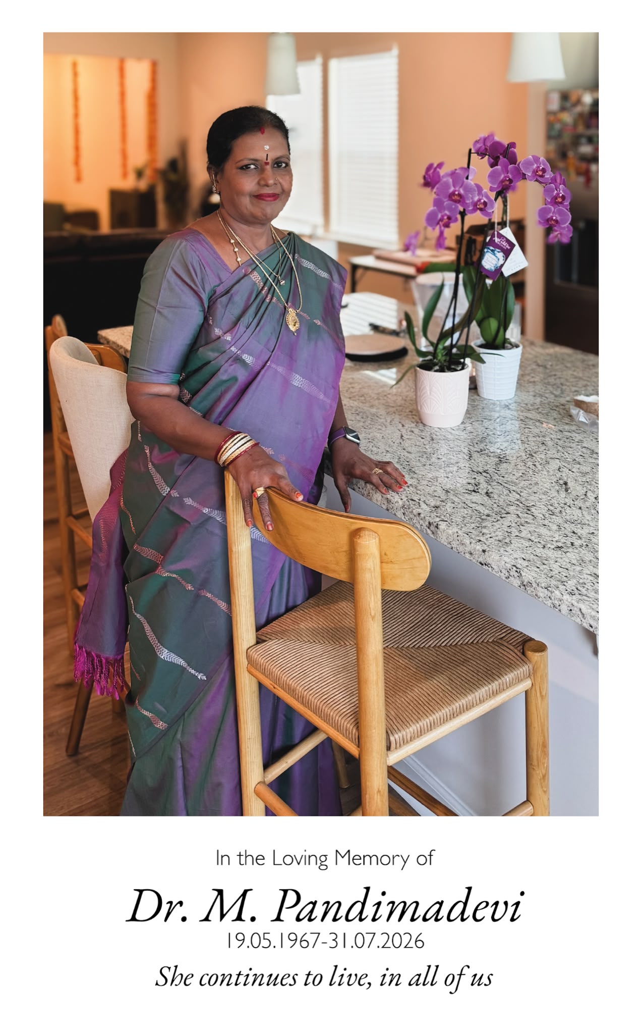 Amma standing by her kitchen counter in a purple and green silk saree, next to orchids. She continues to live, in all of us.