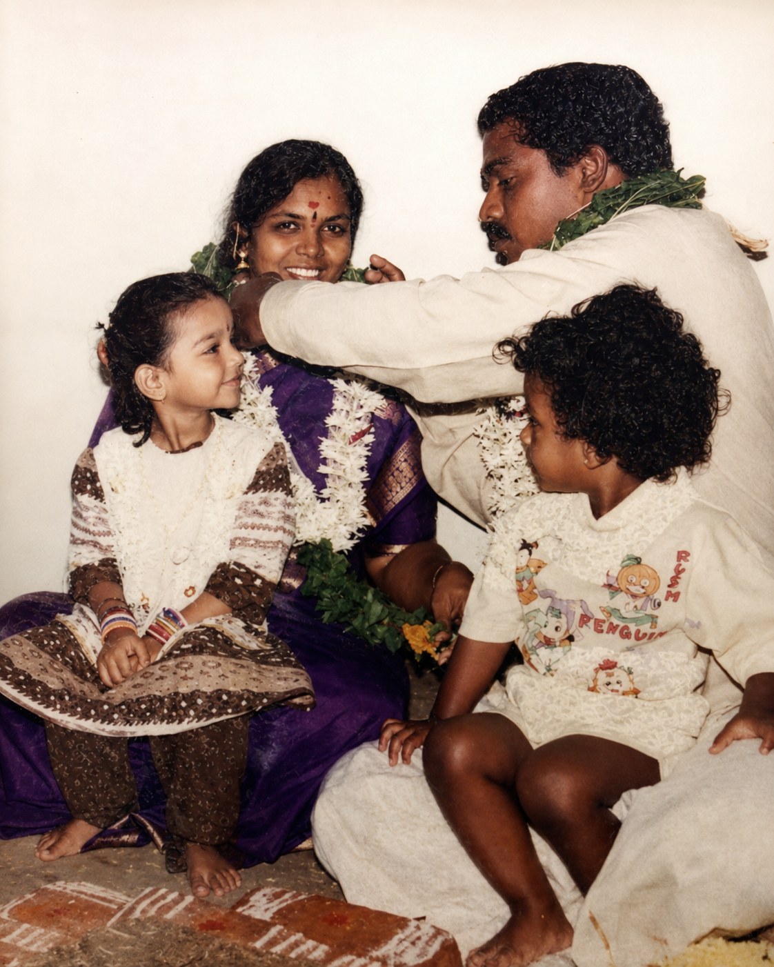 Venkatesan and Devi with their two young children, dressed in traditional clothes with flower garlands, during a family celebration.